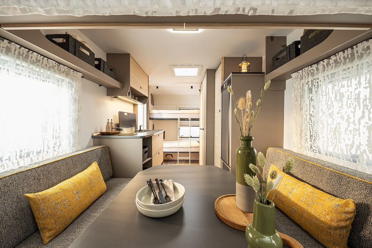 Moderner Innenraum des NEXT Caravan mit grauer Sitzgruppe, gelben Kissen und Blick auf das Etagenbett im Heckbereich